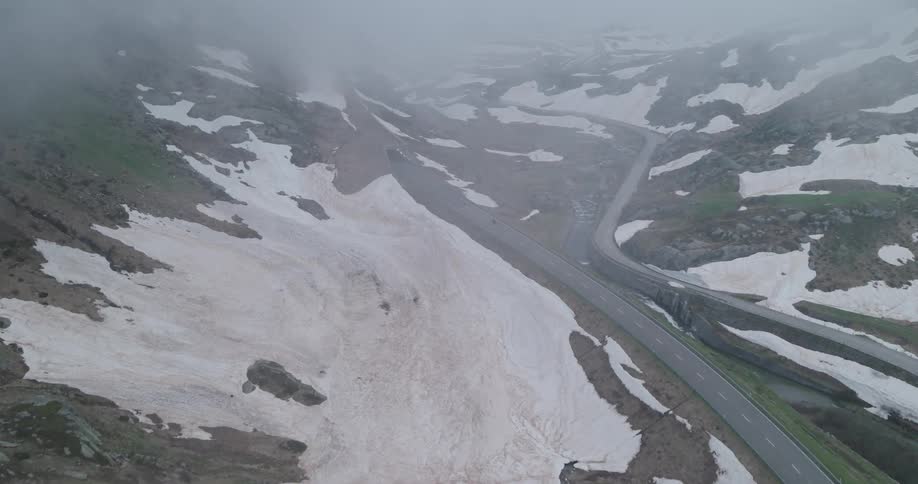 A winding mountain road partially covered in snow stretches into the misty landscape with a lone motorcycle riding into the dark tunnel surrounded by rugged terrain and patches of melting snow