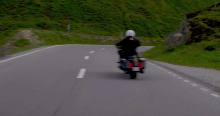 A motorcyclist wearing a helmet rides a touring motorcycle along a winding mountain road with green hills and a stone wall visible in the background