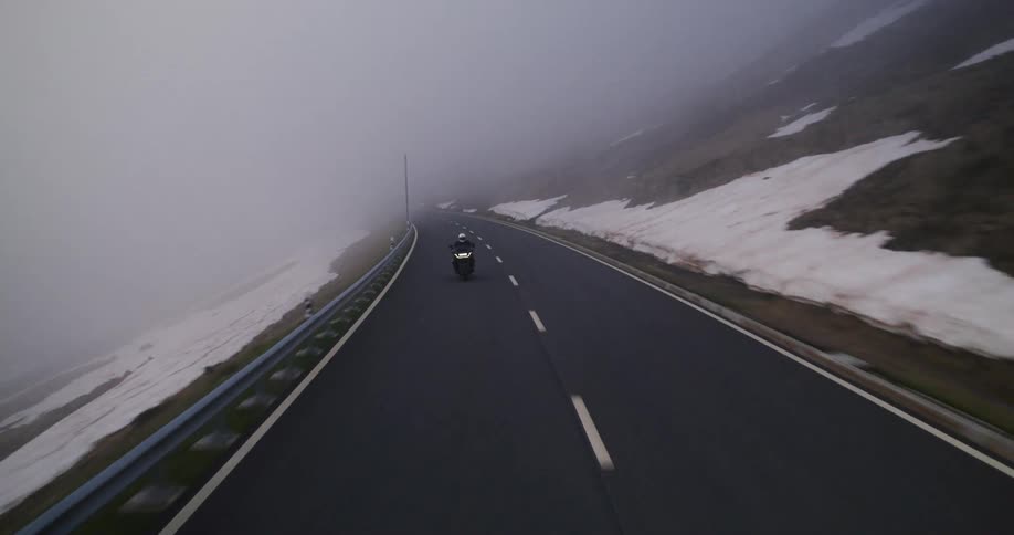 Motorcyclist rides down a mountain road in dense fog with headlights on illuminating the way