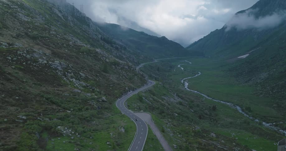 A person rides a motorcycle through a valley at the base of a mountain range on a cloudy day