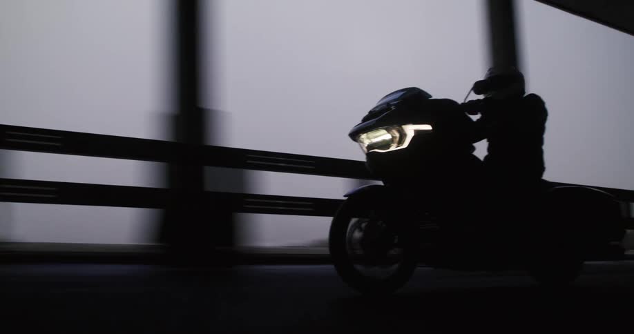 A motorcyclist helmet on and headlights glowing rides smoothly from open road into a dim tunnel navigating past concrete pillars and guardrails on a sleek highway