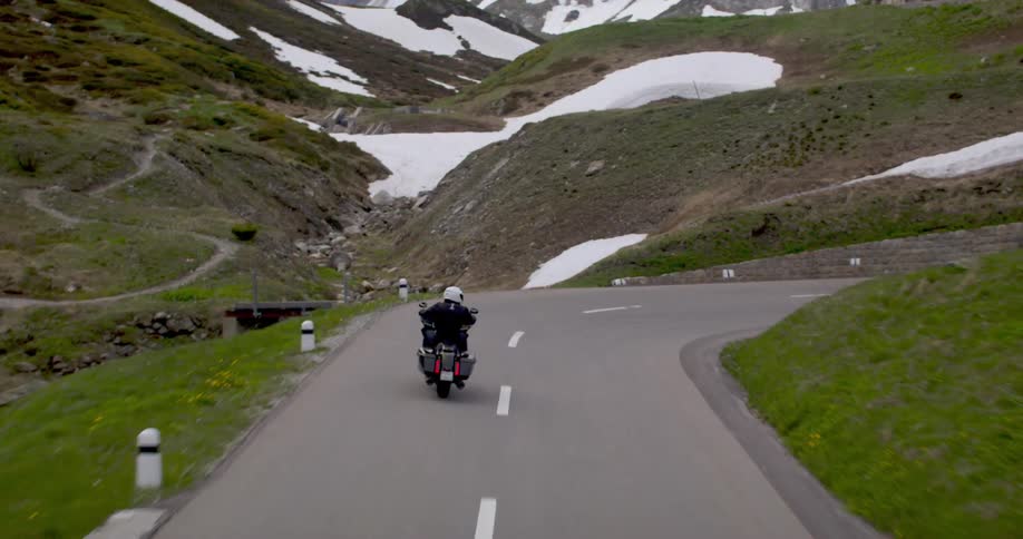 Motorcyclist rides a winding mountain road surrounded by snow and vegetation