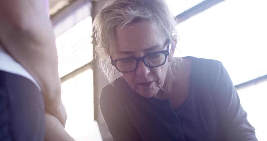 The face of a mature woman wearing glasses who is talking and someone is standing in front of her