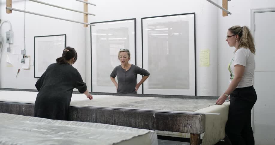Three women collaborate in a bright minimalist workspace carefully working on a large sheet of material spread across a table