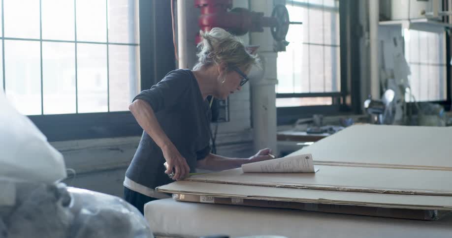 Woman with glasses and tiedup hair analyzes a plan on a large work table in a studio welllit by large windows
