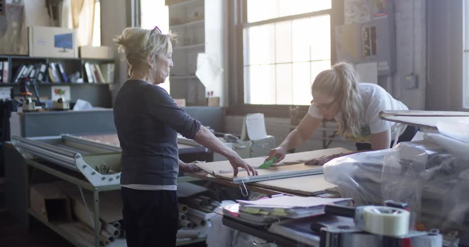 In a sunlit workshop filled with books and tools two women collaborate one holding a measuring tool while the other carefully cuts a piece of material on a large worktable