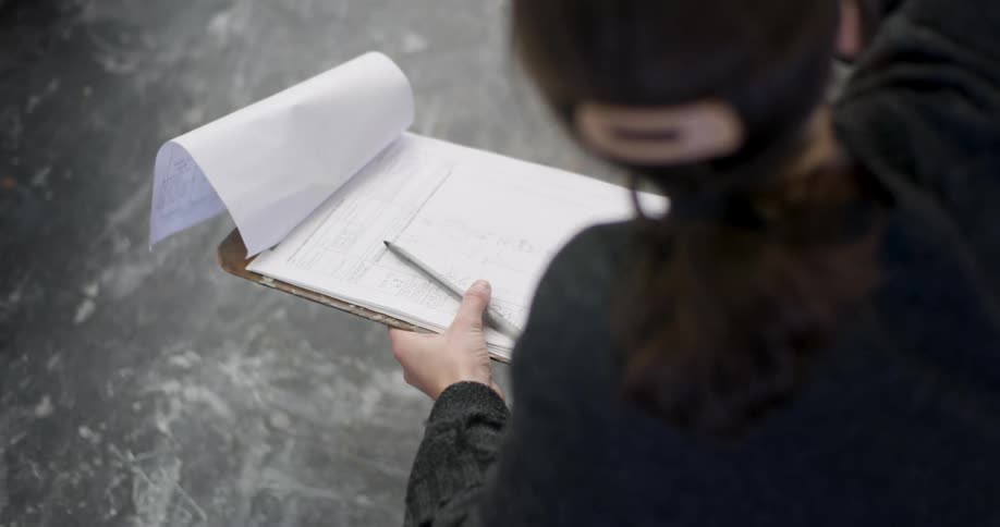 A woman with her hair tied back holds a clipboard flipping a page while signing and talking to a person in front of her