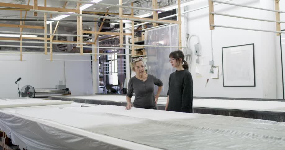 Two women talk in a textile manufacturing workshop with large tables covered in fabrics and wooden structures suspended from the ceiling