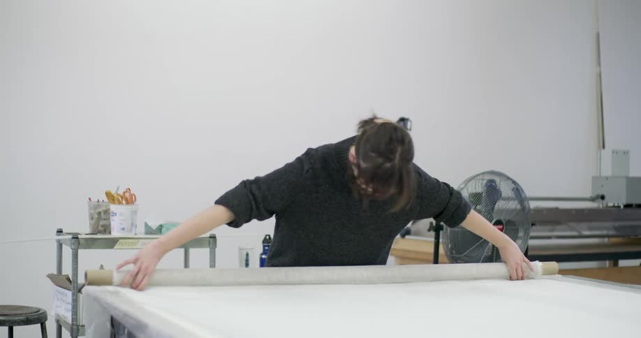 Woman in glasses and a dark sweater unrolls a large roll of fabric on a table
