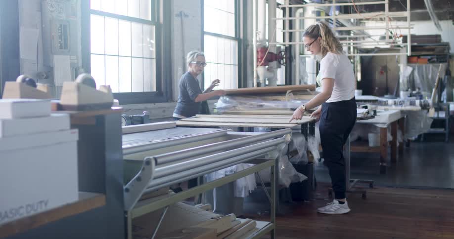 Inside a textile factory two coworker women one younger with dirty blond hair and an older companion are depicted collaboratively lifting a board from a table in a brightly lit indoor space