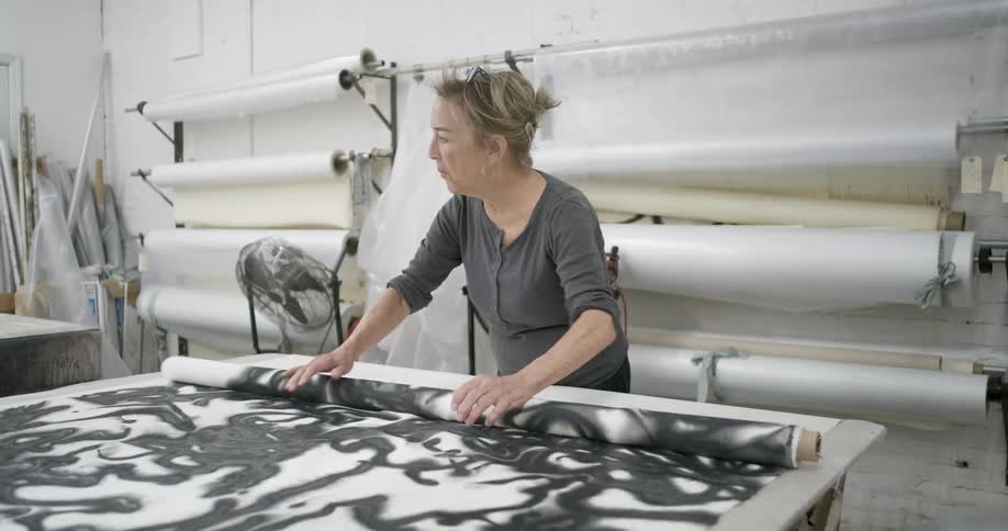 Woman unrolls an abstract patterned fabric in a studio surrounded by rolls of material