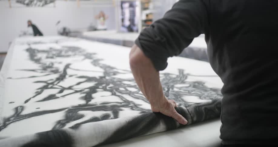 An elderly woman in a dark sweater is rolling out a large piece artwork with black abstract patterns in a bright workspace while others work in the background