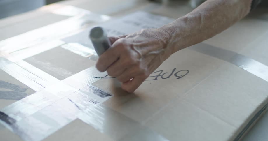 An elderly hand using a thick black marker to write open this on a sealed cardboard box