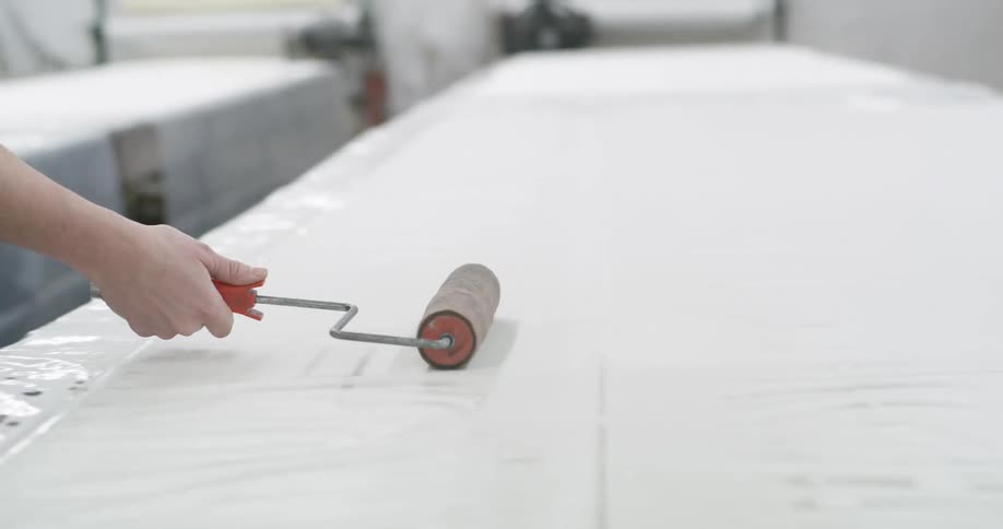 Two women move rollers across the piece of fabric on a long worktable