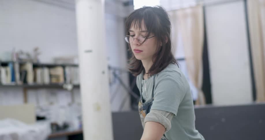 A young woman wearing glasses and a paintstained jumpsuit is crouching down to pick up a roller brush and then running it over a surface in front of her