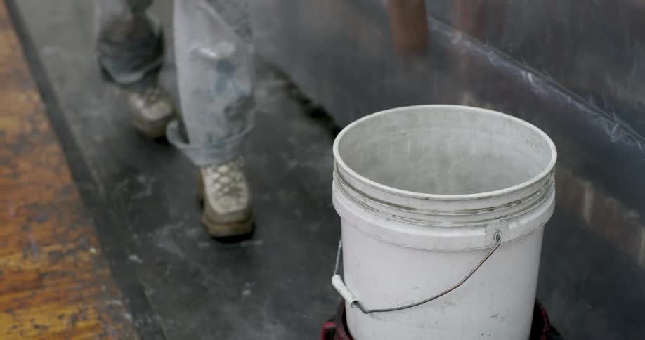 A painter uses a hand roller to pull a bucket which is inside a wheeled cart closer while also wiping the brush inside the bucket