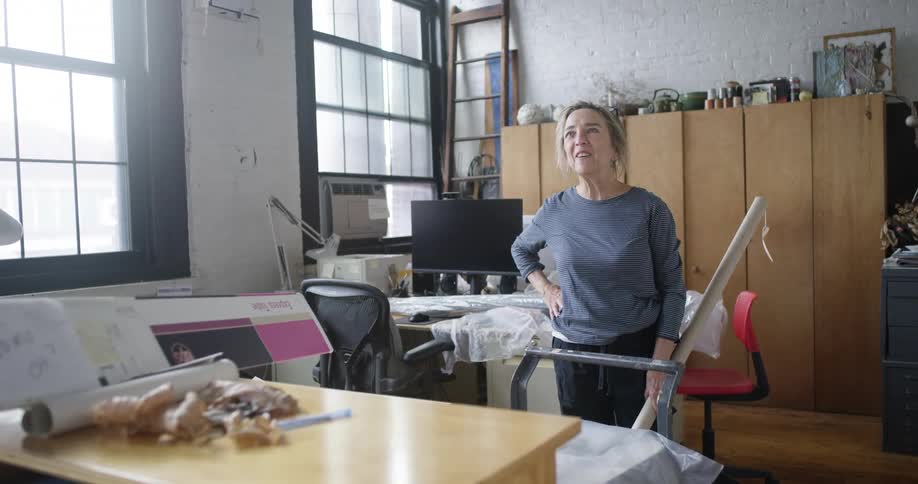 A woman stands in a cluttered office looking upwards with a hopeful expression