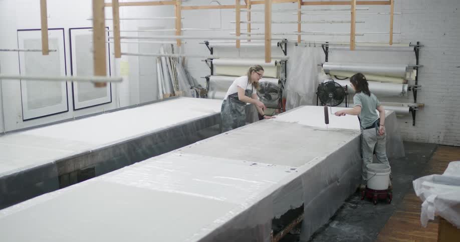 Two coworkers paint a countertop using paint rollers in a large shop