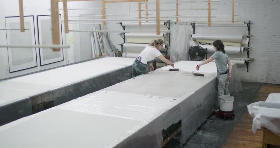 Two women apply paint to large pieces of fabric stretched out on a work table inside a studio