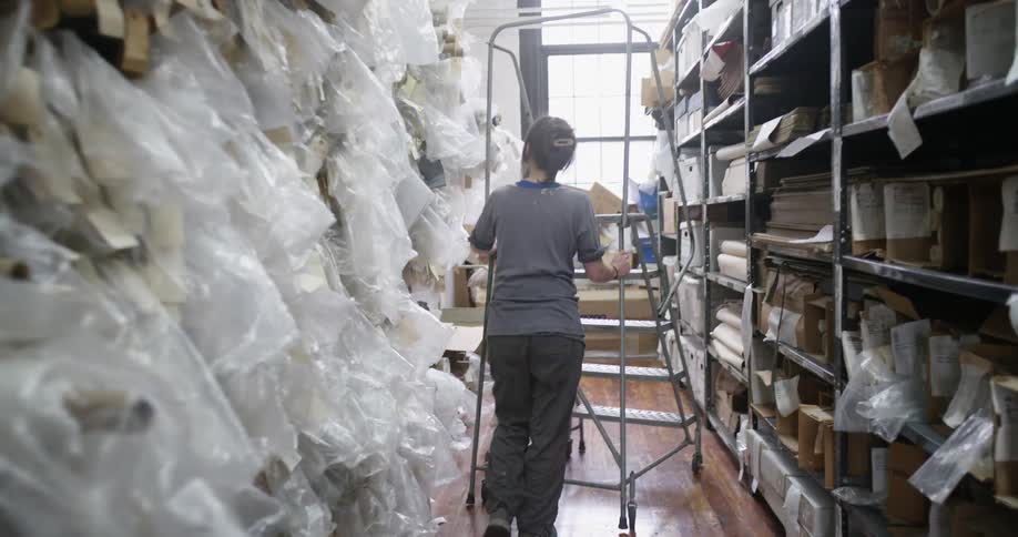 Woman guides a mobile ladder into a filing cabinet crammed with rolls of paper and materials wrapped in plastic