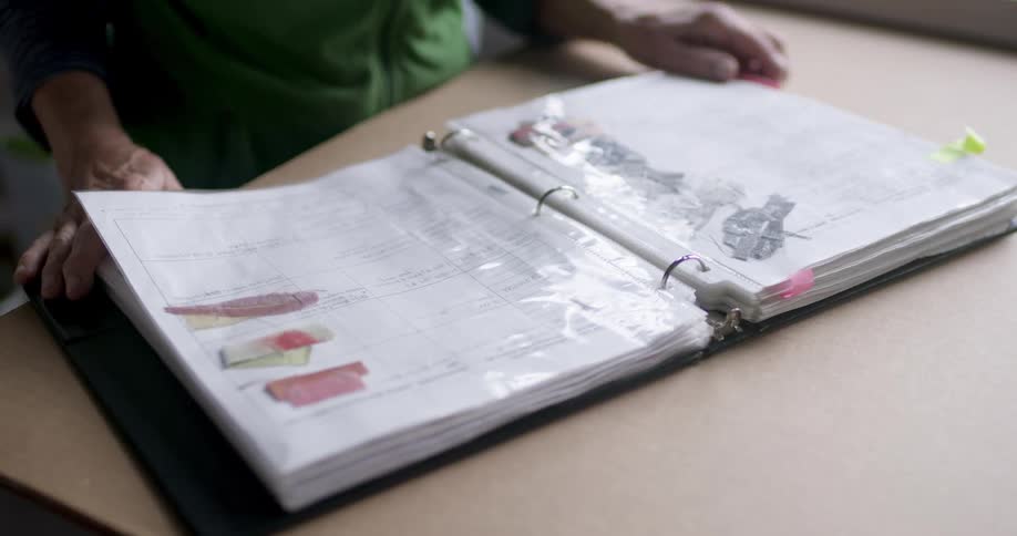 A woman in a green apron is turning the pages of an open binder filled with sketches and color samples which are protected by plastic sleeves