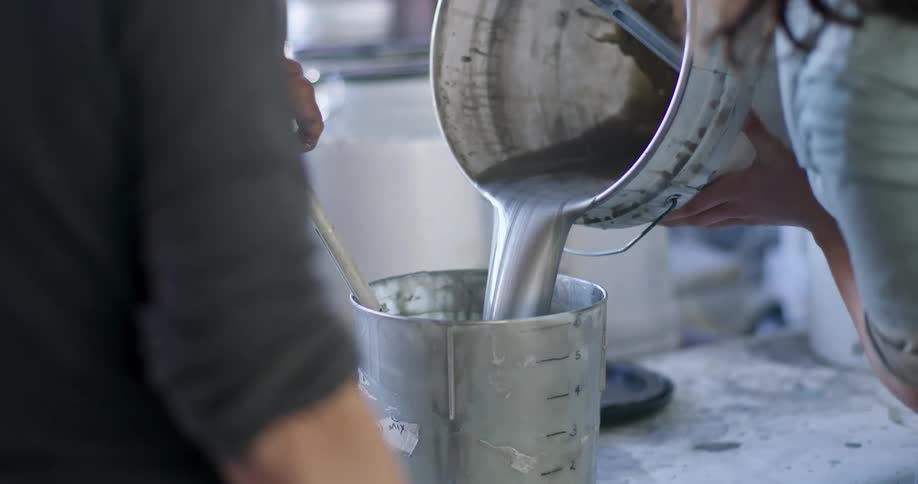 A woman carefully pours a thick metallic silver liquid from a large worn metal bucket into a measuring container while another woman stirs