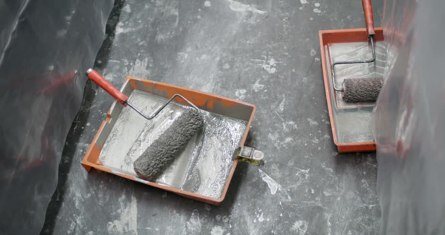 Two paint rollers covered in paint rest in orange trays on a stained floor