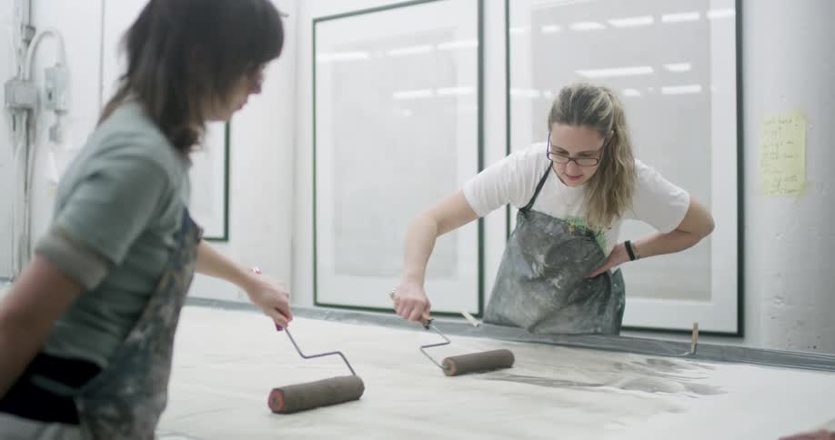Two women wearing aprons work together in an art studio using paint rollers on a large canvas while one leans forward to guide the other surrounded by framed artworks and notes on the wall