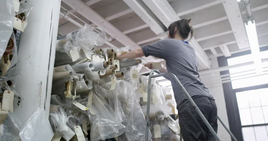 Woman climbs a ladder to retrieve a roll of fabric from an organized storage area inside a studio
