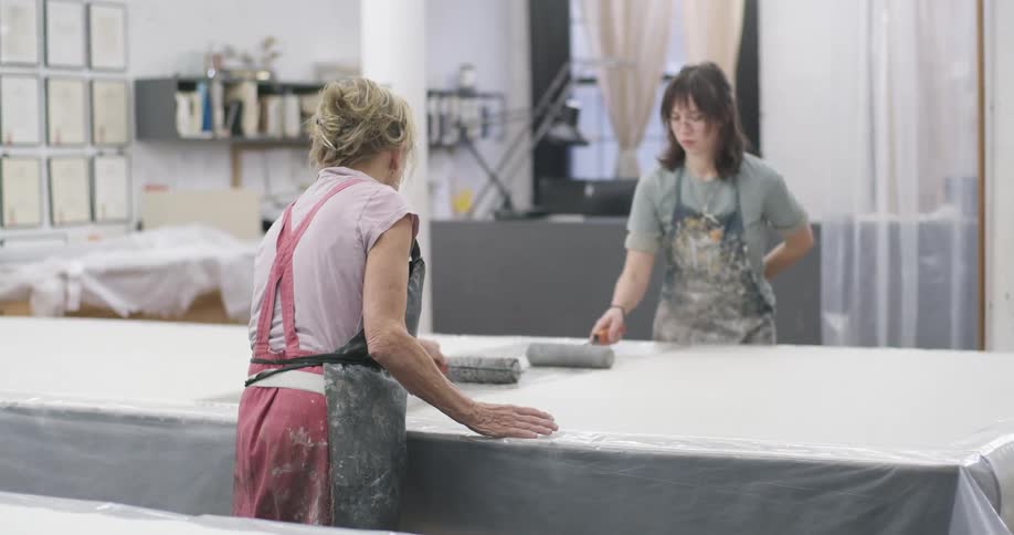 Two women use paint rollers to paint a piece of fabric lying on a plastic sheet covered work table