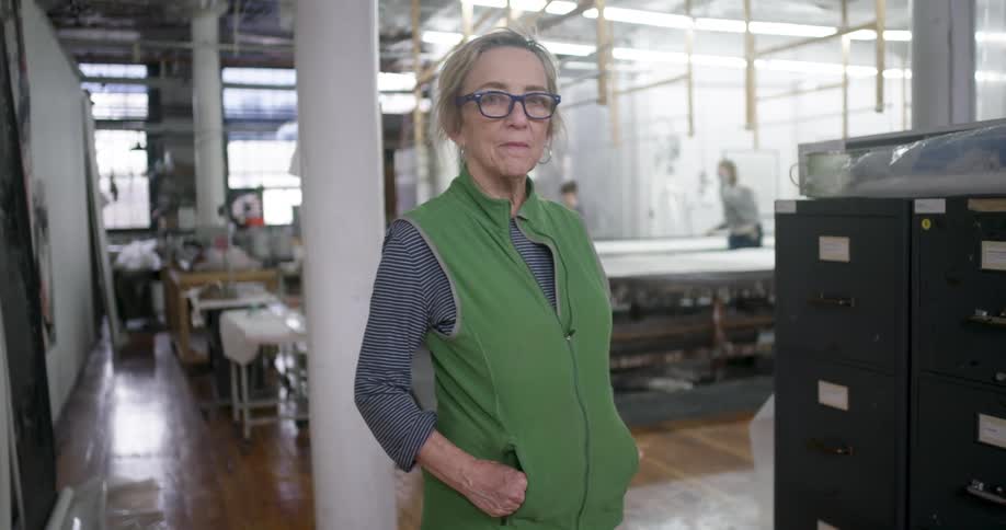 An older lady in a bright green vest confidently poses for a photo in a bustling workshop with the blurred figures of other workers visible in the background as they move between work tables