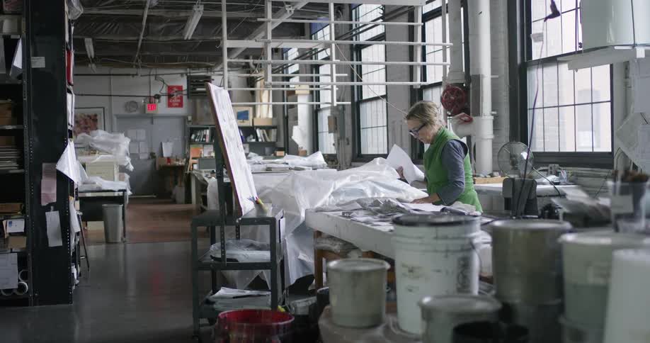 A woman standing wearing a green vest is fiddling with some white fabrics in a factory