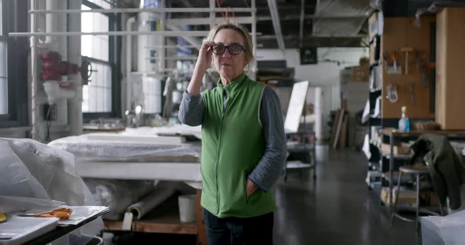 In a quiet textile shop an older lady in a green vest strikes a pose and puts on her glasses becoming the central figure in the otherwise still workspace