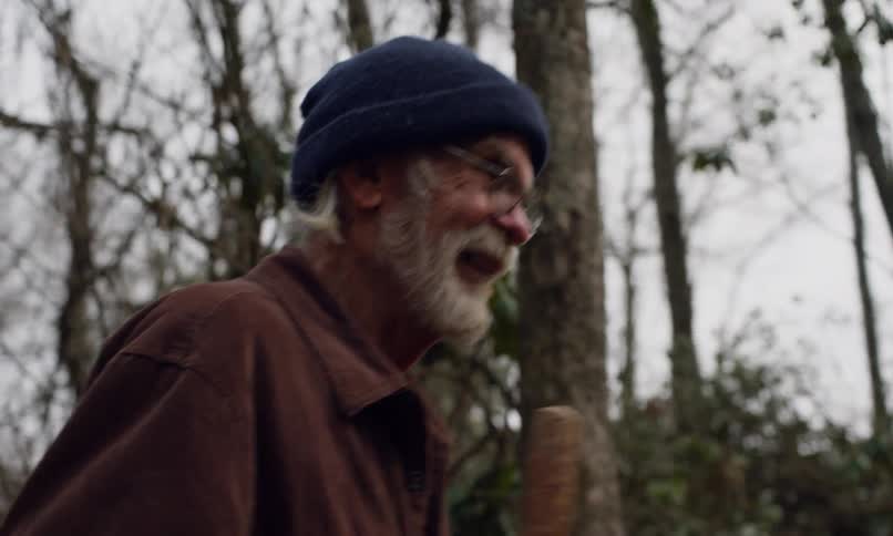 An elderly man with a white beard glasses and a blue beanie walks through a forest holding a stick