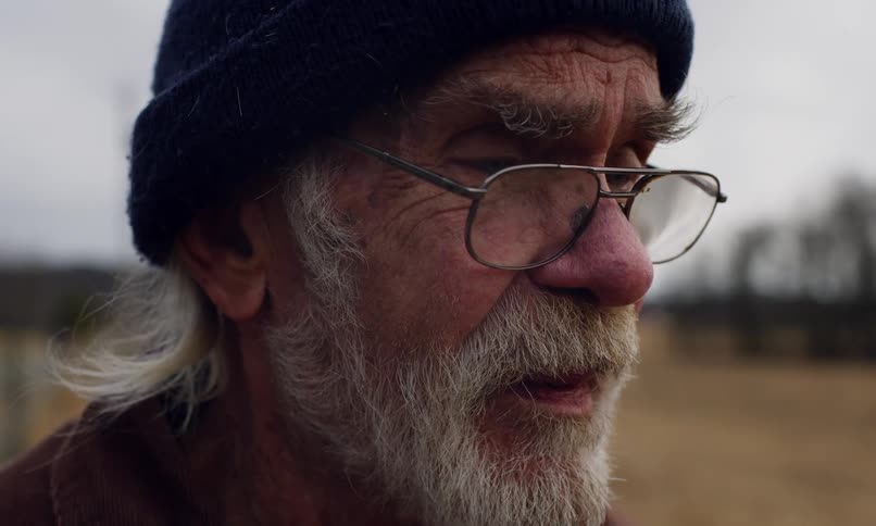 An old man wearing a cap and glasses is smoking a cigarette and blowing smoke out of his mouth