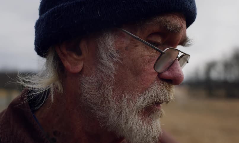 An elderly man with a weathered face white beard and a navy blue beanie gazes downward with a contemplative expression his glasses resting slightly crooked on his nose as he smokes a cigarette