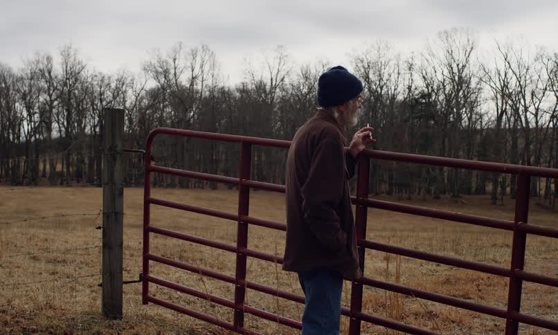 Elderly man in a cap and dark coat smokes while leaning against an iron gate in a dry and cloudy rural landscape