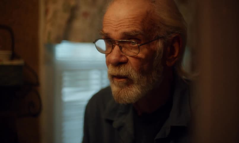 An elderly man with a white beard and glasses sits in a dimly lit room his expression thoughtful as he gazes intently ahead illuminated by the soft glow of natural light from a nearby window