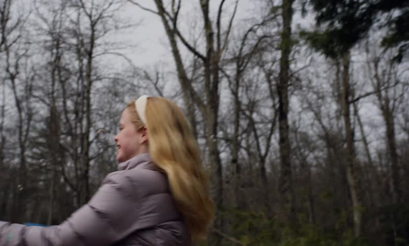 In a playful outdoor scene a young girl joyfully spins in a grassy field operating a blue bubble gun and sending iridescent bubbles into the crisp air as her blond hair swirls with stark bare trees in the background