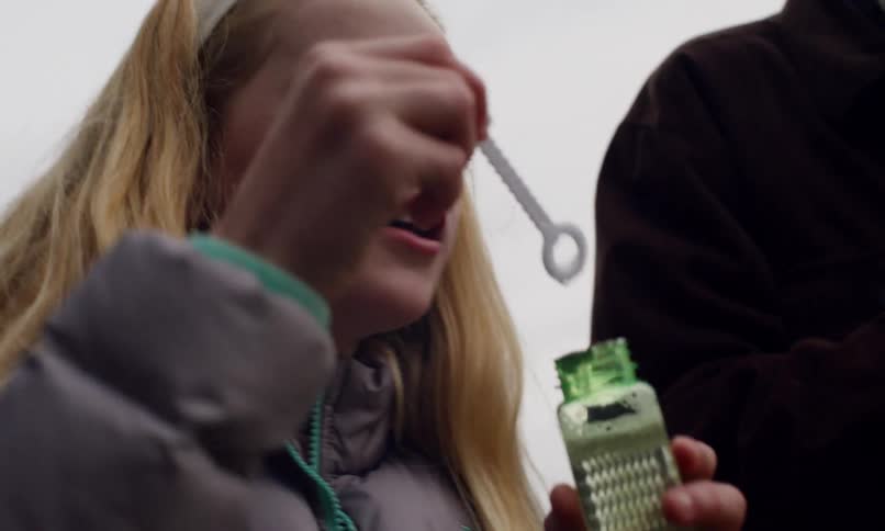 A young girl with long blonde hair blows a bubble through a plastic wand her expression focused while an older man in a dark coat stands beside her partially visible in the frame