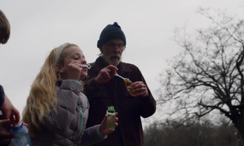 A young girl and an elderly man both bundled up in warm clothing joyfully soap bubbles into the cool outdoor air sharing a playful moment together