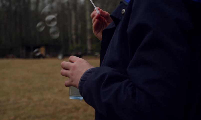 Boy dips a wand into bubble solution then blows bubbles while standing in a yard near a grove of leafless trees