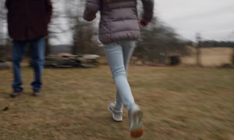 Boy in blue jacket runs ahead of blonde girl in gray coat who chases him with open arms across a grassy field