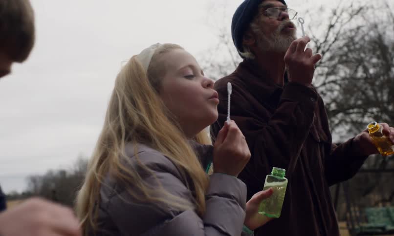 Elderly man in a bonnet and two children soap bubbles outdoors in a cloudy rural setting
