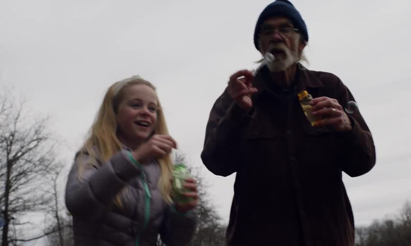 Girl blowing bubbles outdoors with her grandfather and brother chases a bubble to pop it