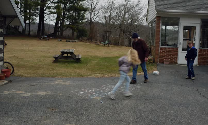 A young girl is playing hopscotch on a chalkdrawn grid while an elderly man and a boy watch her in an outdoor setting near a brick building