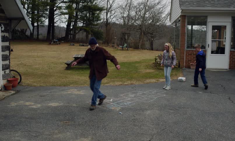 A young girl is playing hopscotch while an elderly man and a boy wait for their turn taking turns in the game on a chalk drawn grid in a peaceful outdoor setting