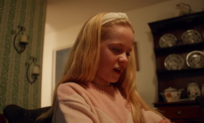 A young girl with blonde hair and a pink sweater sits at a table and plays with dominoes
