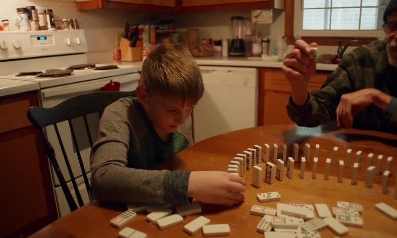 A young boy an elderly man in a beanie and another person gather around a wooden table carefully setting up a circular arrangement of dominoes in a cozy kitchen