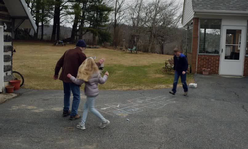 Girl playing hopscotch drawn with chalk on the asphalt while an elderly man and another child watch next to a house in the countryside